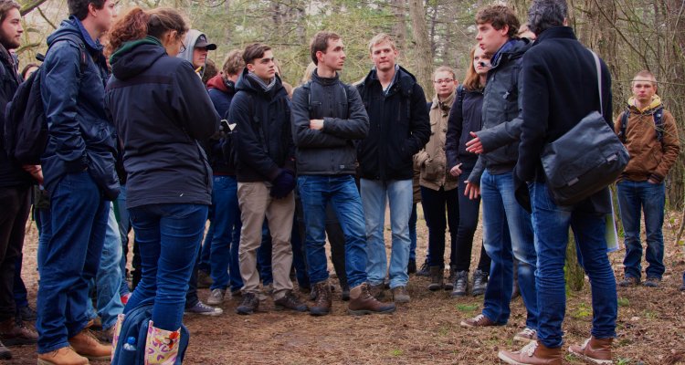 De afbeelding laat een groep mensen in het bos zien. Het ziet eruit alsof iemand iets uitlegt aan de rest.